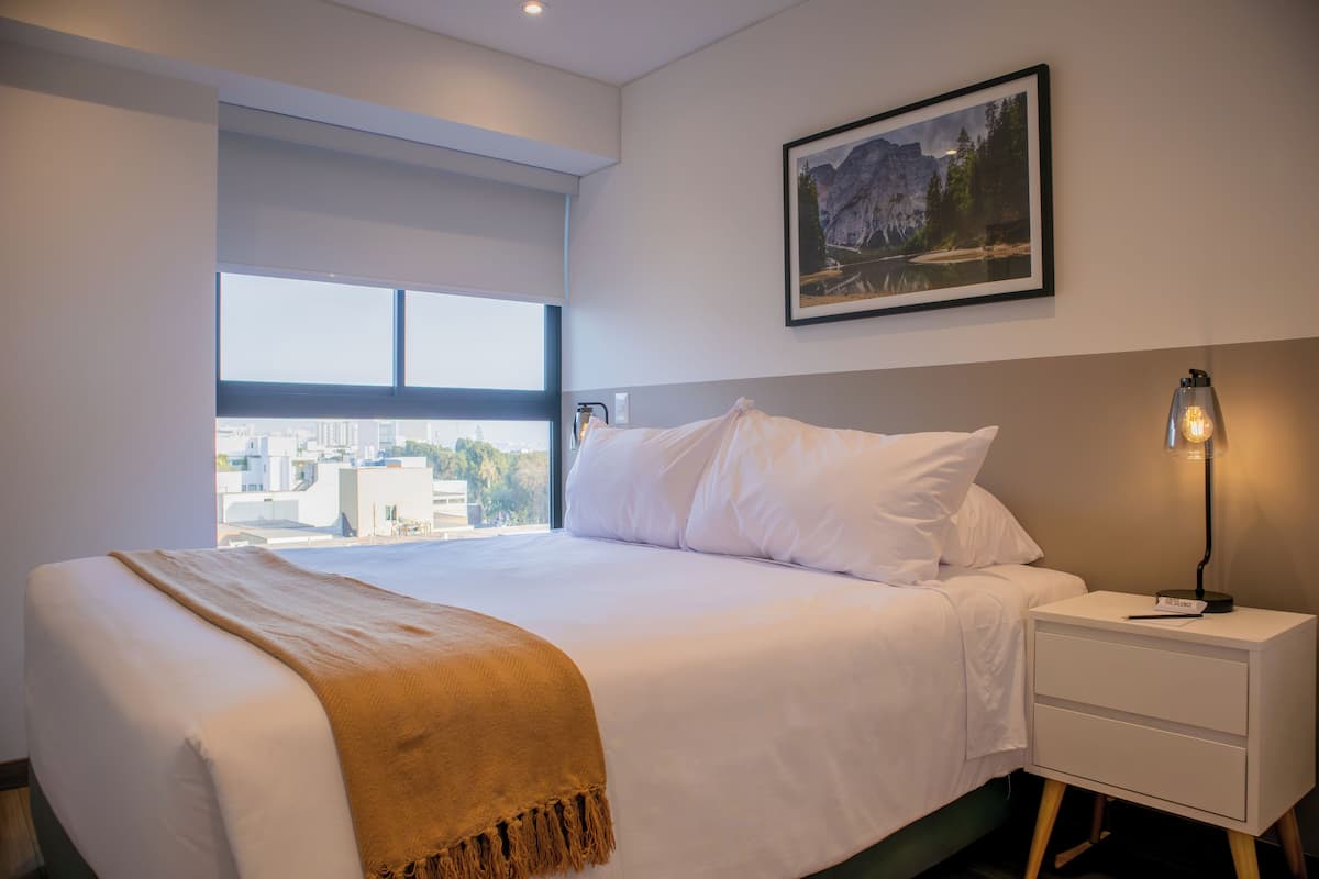 A modern hotel room with a large bed, a nightstand, a lamp, and a framed picture on the wall.