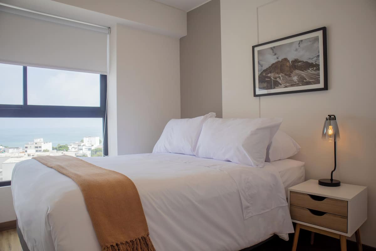 A bedroom with a bed, a nightstand, a lamp, a framed picture, and a window with a city view.