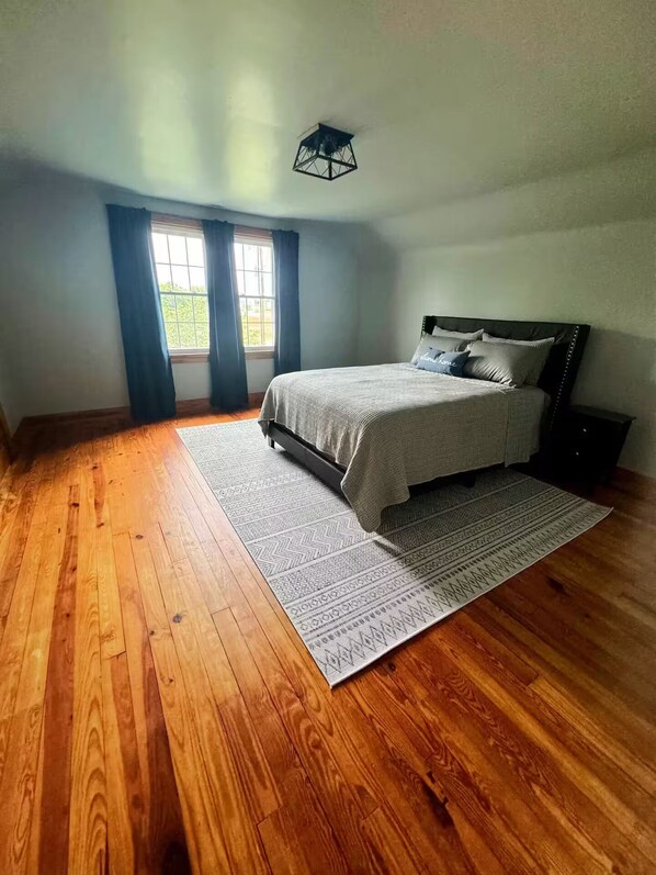 A bedroom with a wooden floor, a bed with a grey bedspread, a window with blue curtains, and a ceiling light fixture.