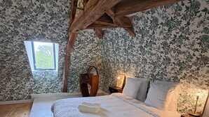 Un dormitorio con techo de madera, papel tapiz floral, una cama con ropa blanca, una mesita de noche con lámpara y una ventana que da a zonas verdes.