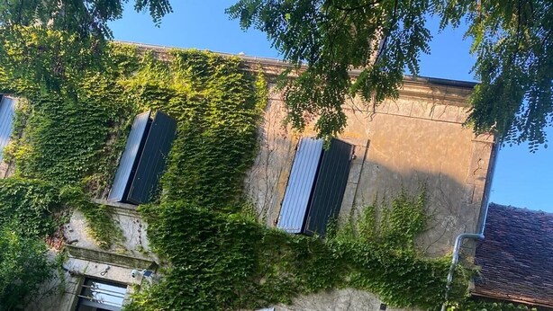 Una mesa de comedor rústica al aire libre con una botella de vino, copas y platos de comida, frente a un edificio de piedra con hiedra y contraventanas.