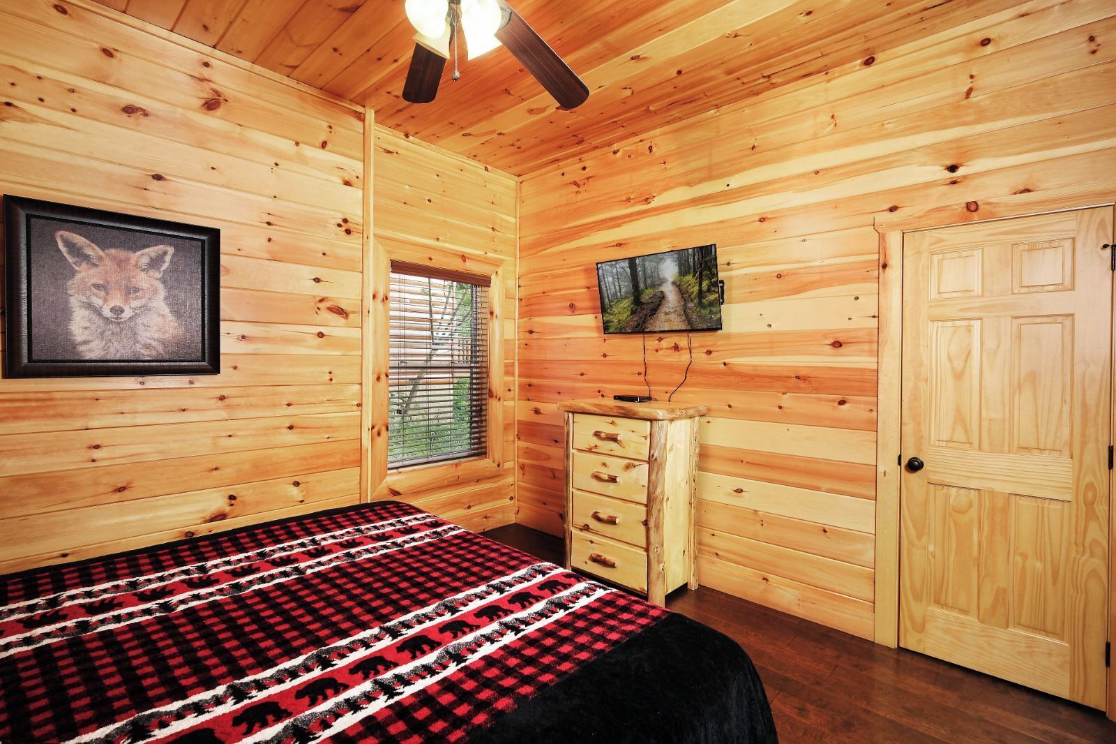 A wooden cabin bedroom with a bed, dresser, TV, and framed picture.