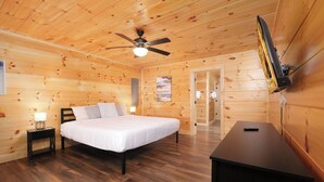 A wooden cabin room with a bed, a desk, a ceiling fan, and a door leading to another room.