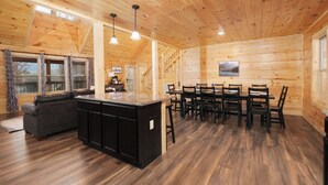 A spacious wooden cabin with a large dining area, a kitchen with a central island, and a cozy living area with a sofa and a fireplace.