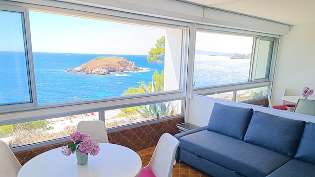 A room with a large window overlooking a rocky coastline and the sea. There is a grey sofa, a white table with pink cushions, and white chairs. - Exceptional sea view, air conditioning, parking, private beaches, swimming pools, tennis court, marina. (Bandol)