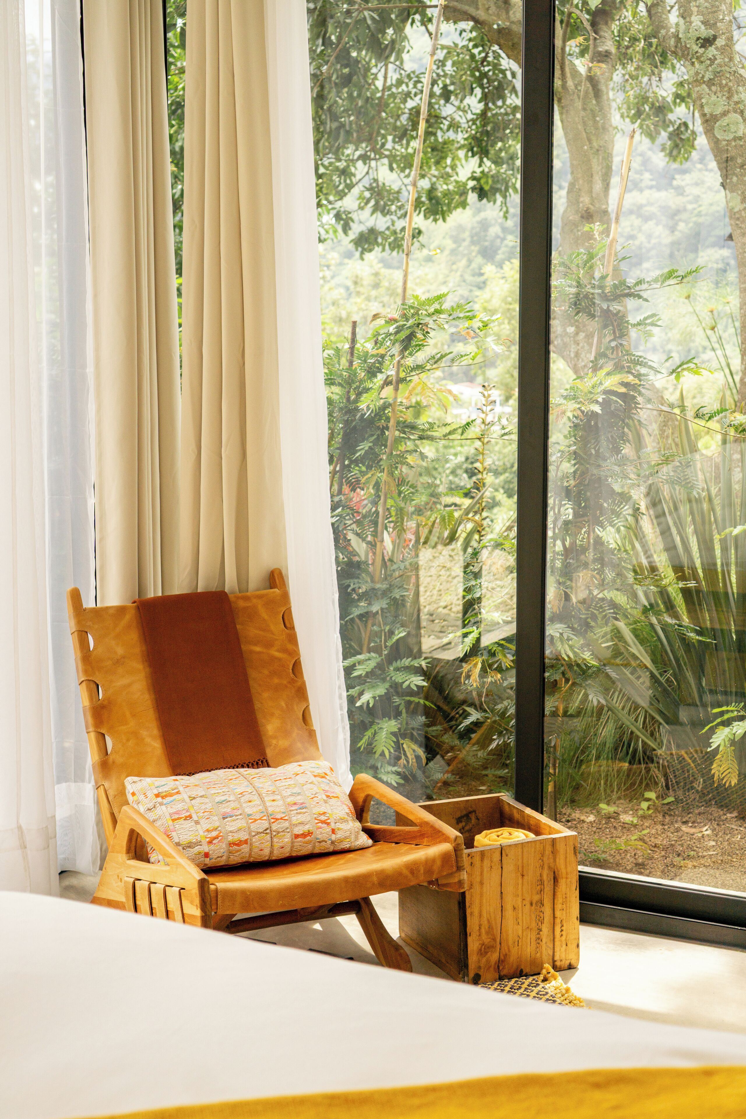 A wooden chair with a cushion and a small wooden box, positioned near a window with sheer curtains, overlooking a lush green garden.