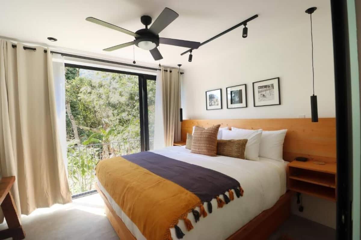 Een slaapkamer met een groot bed, een plafondventilator en uitzicht op bomen door een schuifdeur.