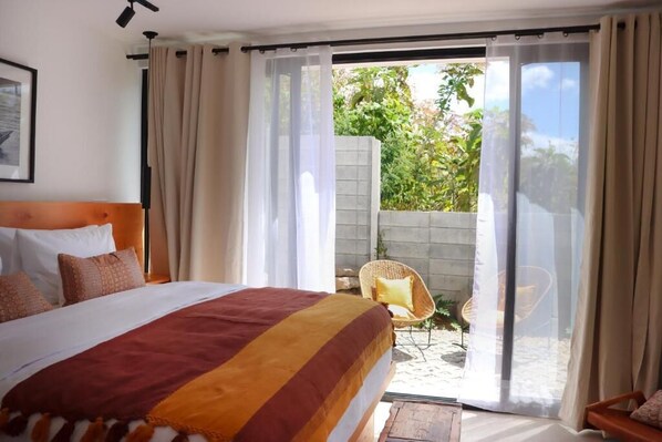 A bedroom with a large bed, a view of a garden through a sliding glass door, and a wicker chair outside.