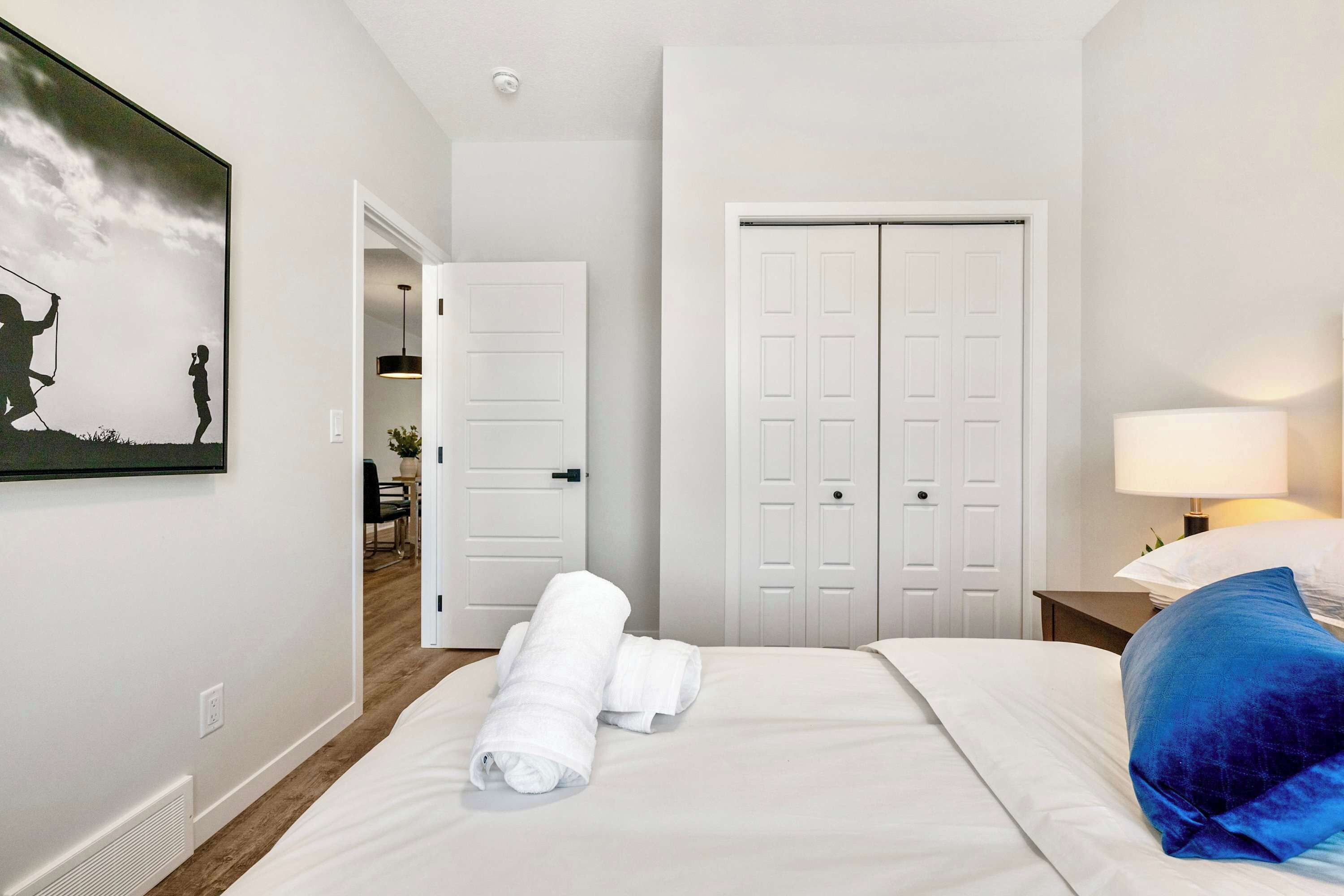 Een hotelkamer met een bed, witte handdoeken, een blauw kussen, een schilderij aan de muur en een deur naar een andere kamer.