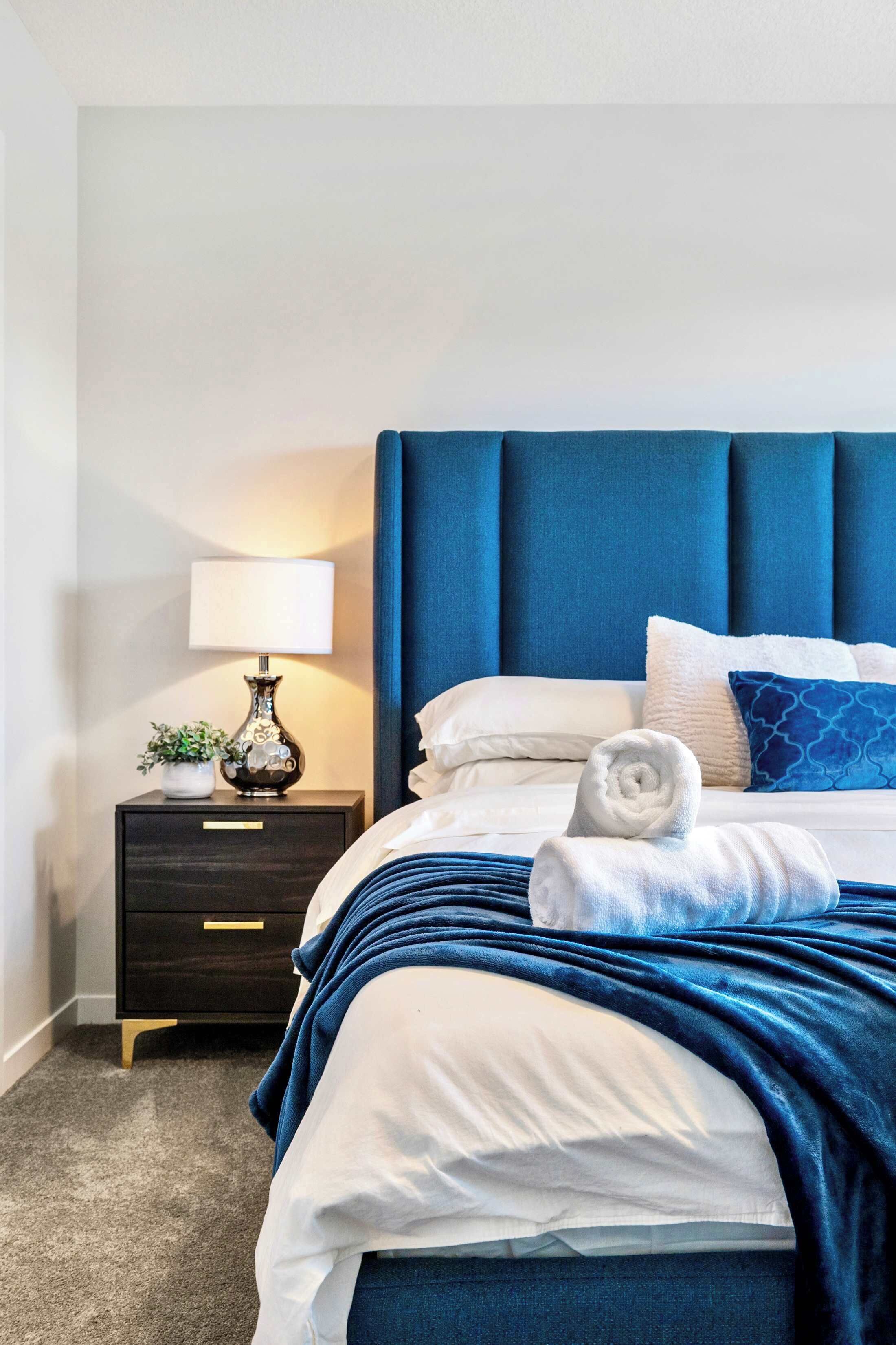 A neatly made bed with a blue headboard, a bedside lamp, and a small plant on a nightstand.