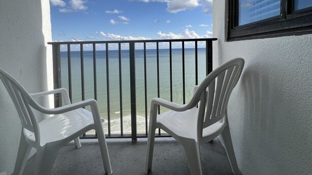 Un balcone con due sedie di plastica bianca, una ringhiera nera e vista sull'oceano sotto un cielo parzialmente nuvoloso.