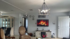 A living room with a fireplace, a television mounted above it, and a chandelier. - Charming Country Home, Pool, Pet Friendly (Clewiston)
