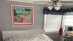 Un dormitorio con una cama cubierta por una colcha de estampado geométrico, un ventilador de techo, una ventana con persianas y un cuadro en la pared.