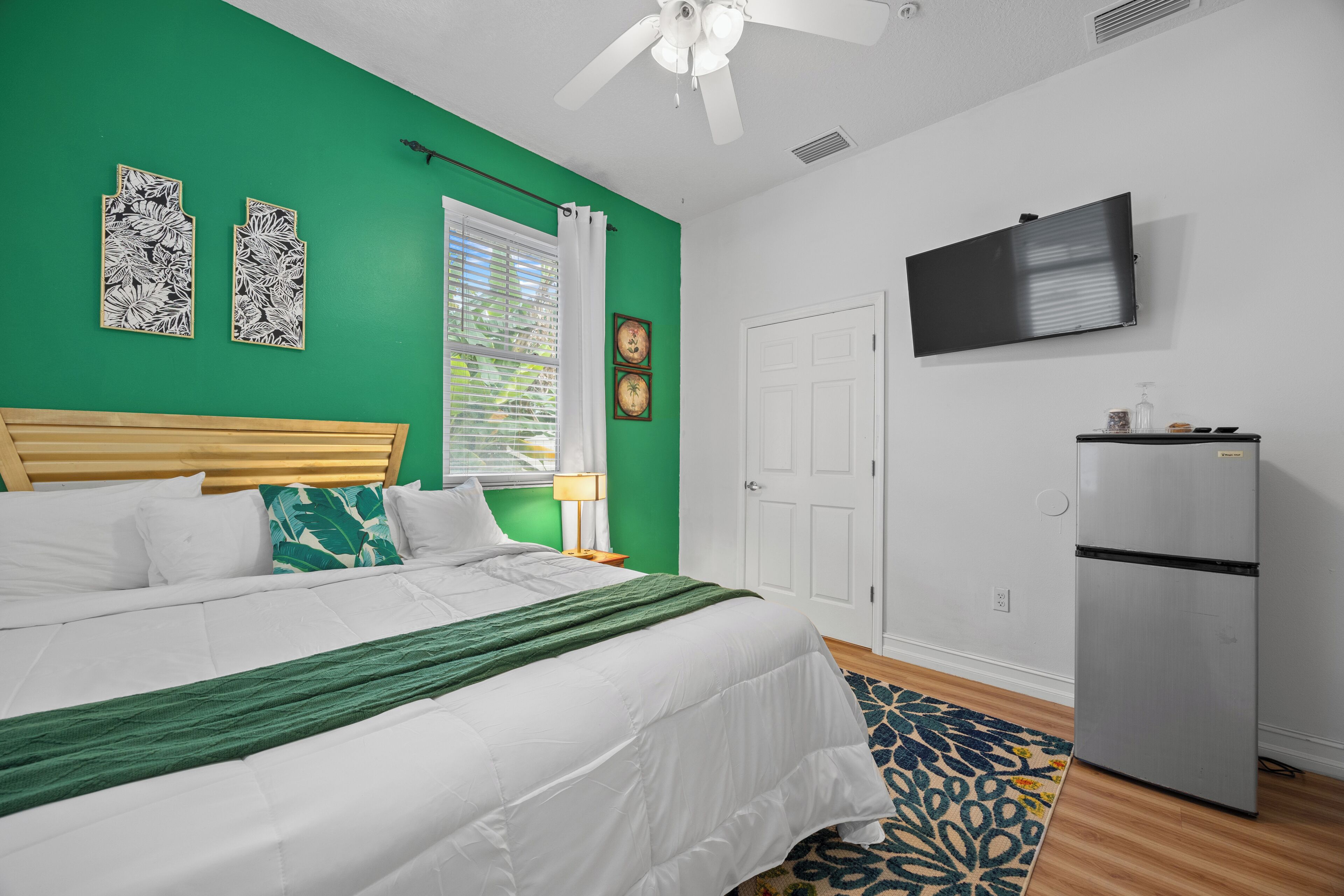 A bedroom with a bed, a ceiling fan, a TV, a refrigerator, and a window with curtains.