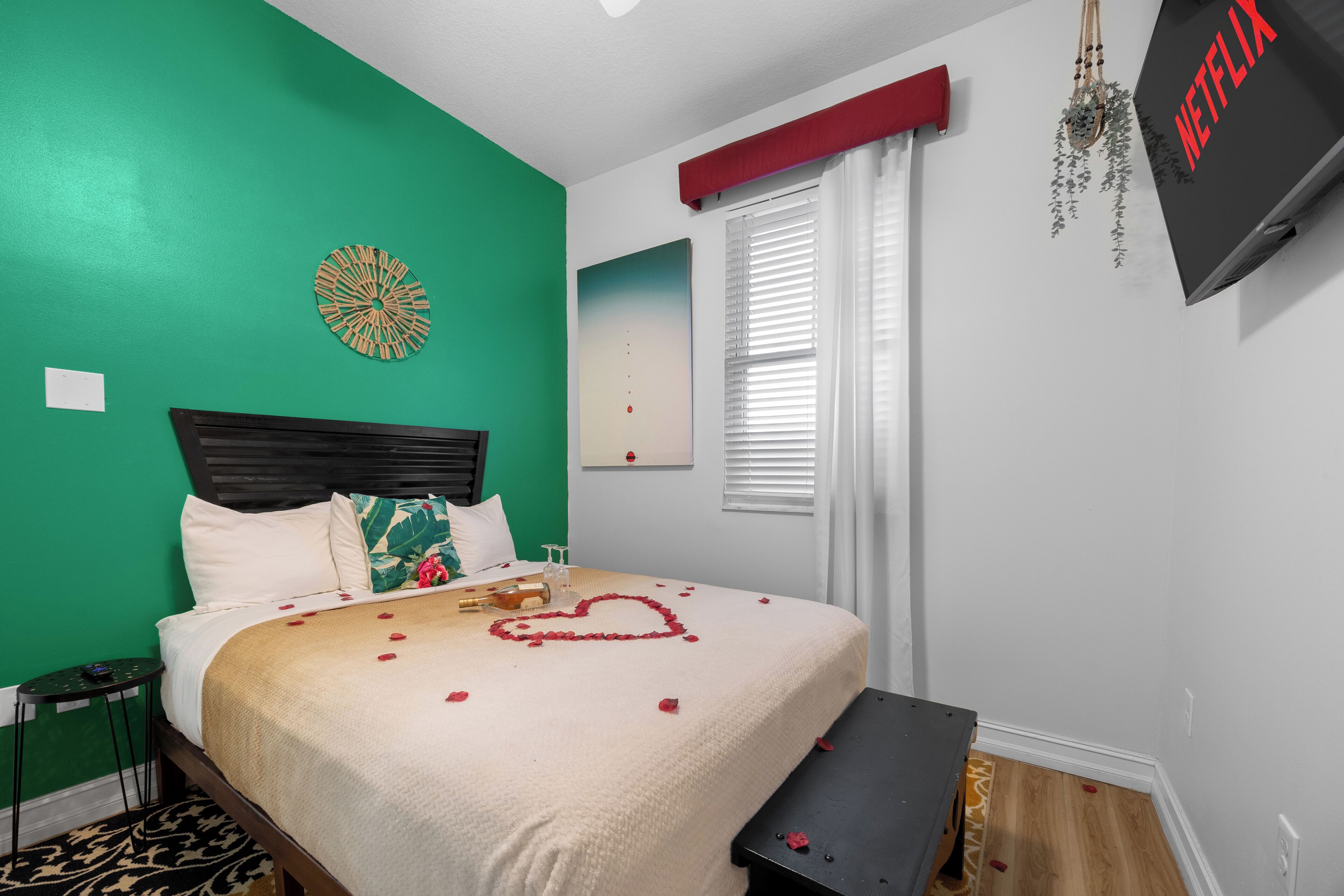 A neatly made bed with a heart-shaped decoration, a wall clock, and a TV on a stand.