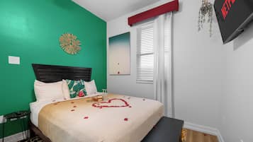 A neatly made bed with a heart-shaped decoration, a wall clock, and a TV on a stand.