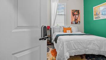 Un dormitorio con cama, mesita de noche, ventana con cortinas, un cuadro en la pared y una puerta.