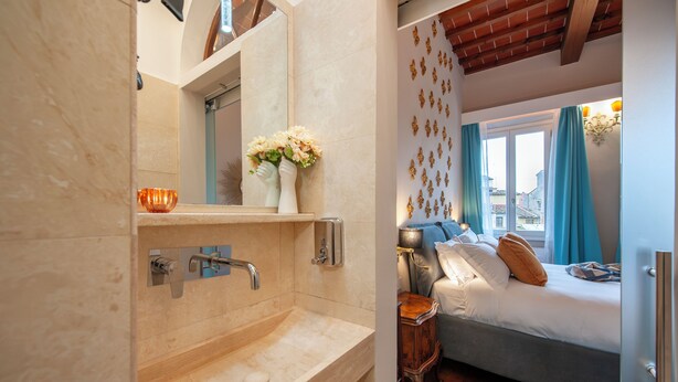 Ein modernes Badezimmer mit einem beigefarbenen Waschbecken, einem Spiegel und einem kleinen Fenster. Neben dem Badezimmer befindet sich ein Schlafzimmer mit einem Bett, Kissen und einem Nachttisch.