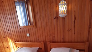 A wooden room with two beds, each with white bedding and a striped blanket. There is a window with a curtain, and a wall-mounted lantern.