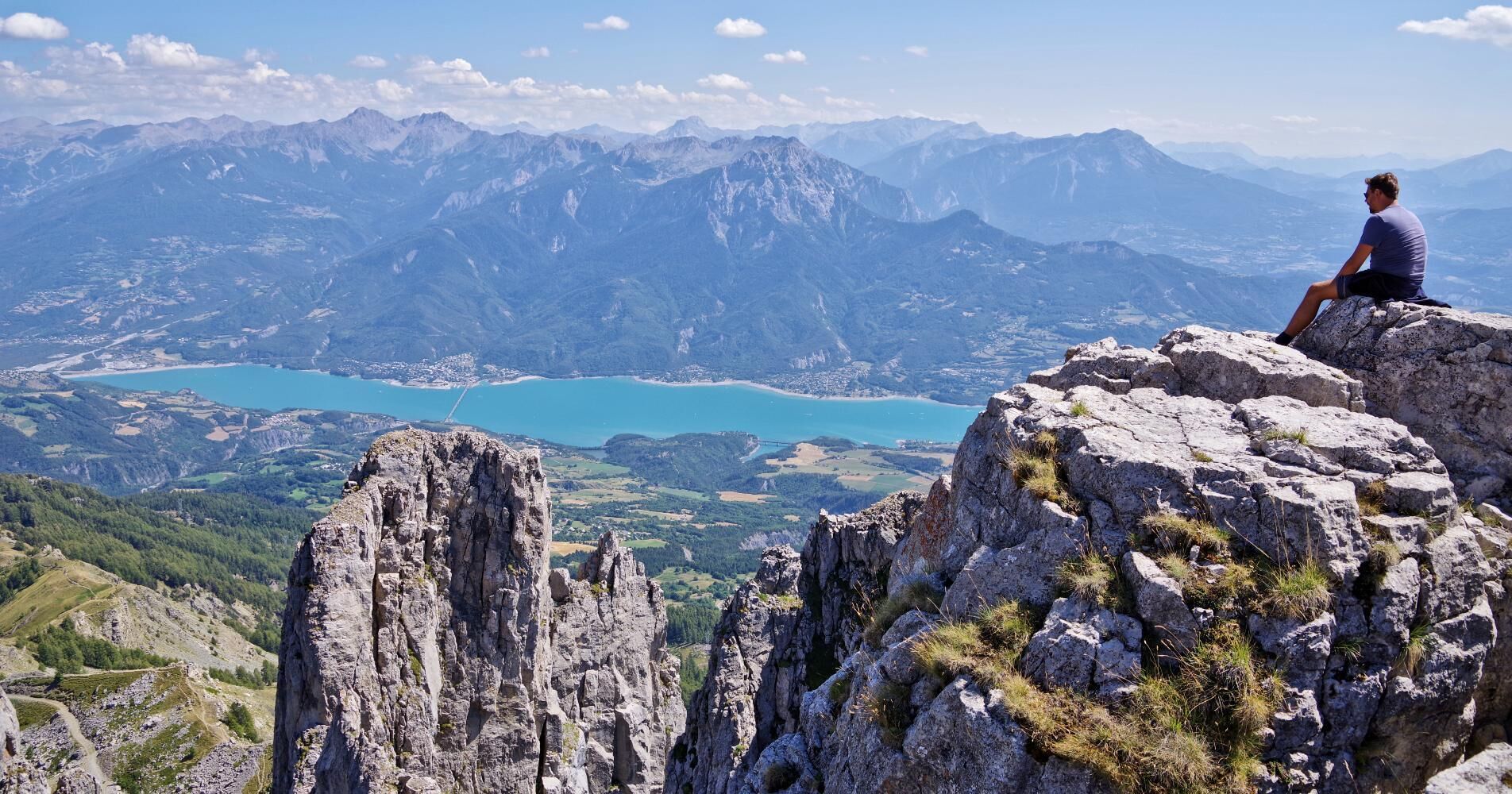 Uma pessoa sentada em um afloramento rochoso, com vista para uma paisagem montanhosa e um lago.