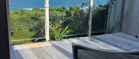 Una terraza de madera con una silla metálica, con vista al mar, vegetación y cielo despejado.