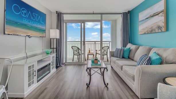 A modern living room with a large TV, a sofa, a coffee table, and a balcony with chairs. - Camelot Getaway Renovated in 2025 (Myrtle Beach)