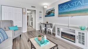 A modern living room with a fireplace, a dining area, and a hallway leading to another room. - Camelot Getaway Renovated in 2025 (Myrtle Beach)