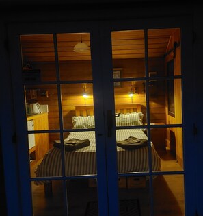 A room with a bed, a desk, and a chair. There is a window with a view of another room.