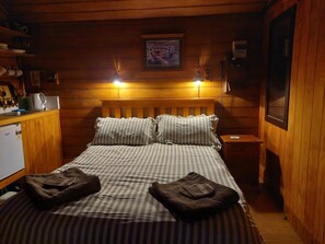 A cozy wooden cabin room with a bed, two bedside lamps, a small table, and a refrigerator.