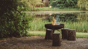 A campfire with logs around it, set in a natural outdoor area with a pond and greenery in the background. - Cosy Cabin in Idyllic Woodland Setting (Storwood)