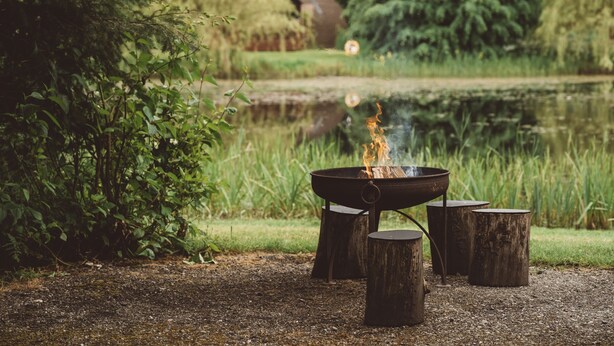 A campfire with logs around it, set in a natural outdoor area with a pond and greenery in the background. - Cosy Cabin in Idyllic Woodland Setting (Storwood)