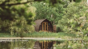 A wooden cabin with a glass door, surrounded by dense greenery and a calm lake. - Cosy Cabin in Idyllic Woodland Setting (Storwood)