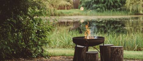 A campfire with logs around it, set in a natural outdoor area with a pond and greenery in the background.