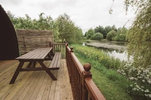 Een houten terras met een picknicktafel en een leuning, met uitzicht op een vijver omringd door groen.