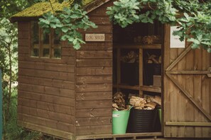 Een houten blokkenberging met een bord waarop 'LOG SHED' staat. Binnenin staan planken met houtblokken en een groene prullenbak.