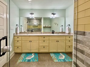 A bathroom with a double sink, beige cabinets, a granite countertop, and a mirror above the sink. There are two towels hanging on towel bars and a bird-themed rug on the floor. - Red Drum 5/4.5 With Pool and Hot Tub (Port O'Connor)