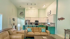 A spacious living room with a brown leather sofa, a matching armchair, and a coffee table. A kitchen with white cabinets and a dining area with a green table and chairs is visible in the background.