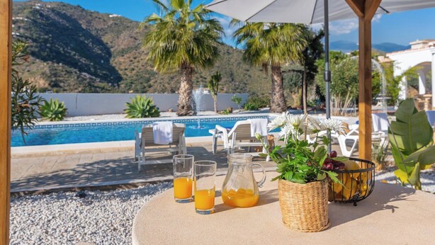 A poolside table with drinks and a basket of fruit, overlooking a pool and mountains.