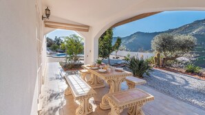 An outdoor patio with ornate tables and benches, a pool in the background, and a mountainous landscape view.