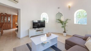 A modern living room with a flat-screen TV on a white console, a brown sofa, a coffee table with books, and a potted plant.