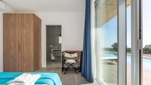 Een moderne hotelkamer met een bed, een houten garderobe, een bureau met stoel en een groot raam met uitzicht op een zwembad.