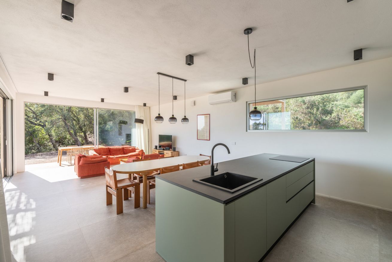 Une cuisine moderne dotée d’un îlot central, d’un coin repas avec une table et des chaises en bois, et d’une grande fenêtre offrant une vue sur les arbres.