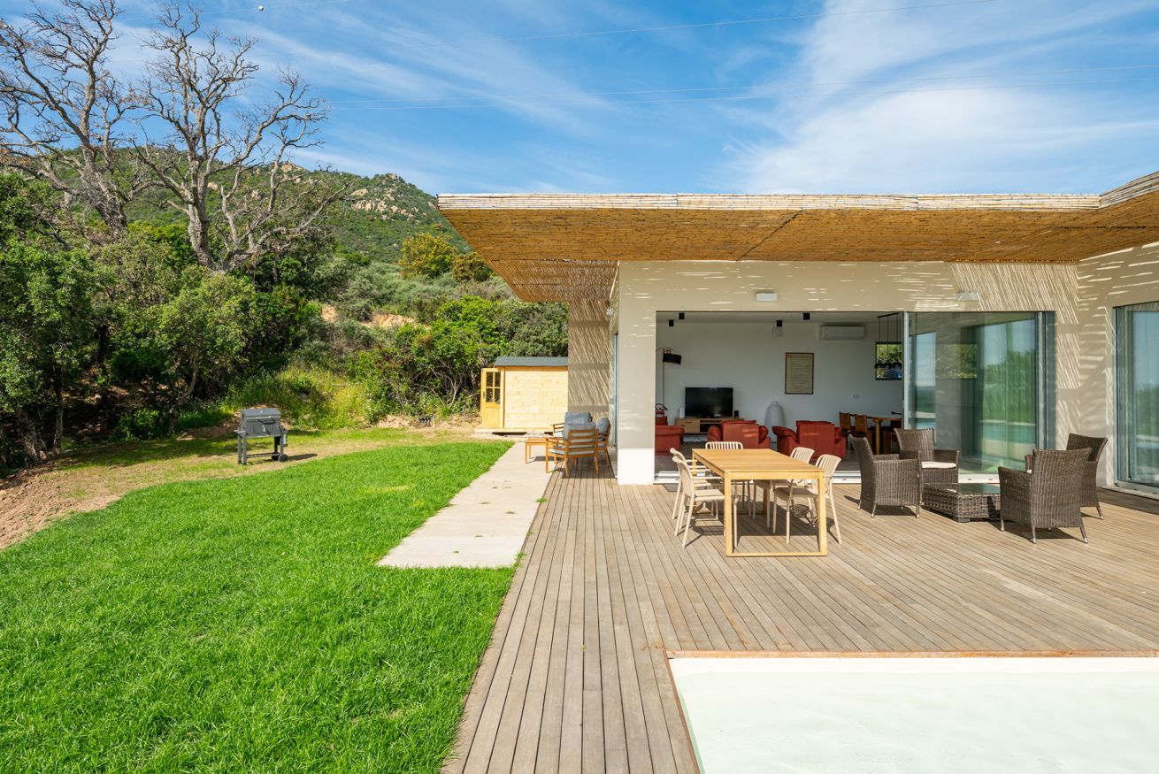 Een modern terras buiten met een houten terras, zithoek en een glazen kamer met een open haard.