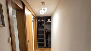 Um corredor estreito com piso de madeira, uma porta à esquerda e um armário com prateleiras e um aspirador de pó à direita.