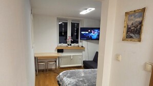 Corredor estreito que dá para um quarto, com uma cama, uma escrivaninha com computador e uma televisão.
