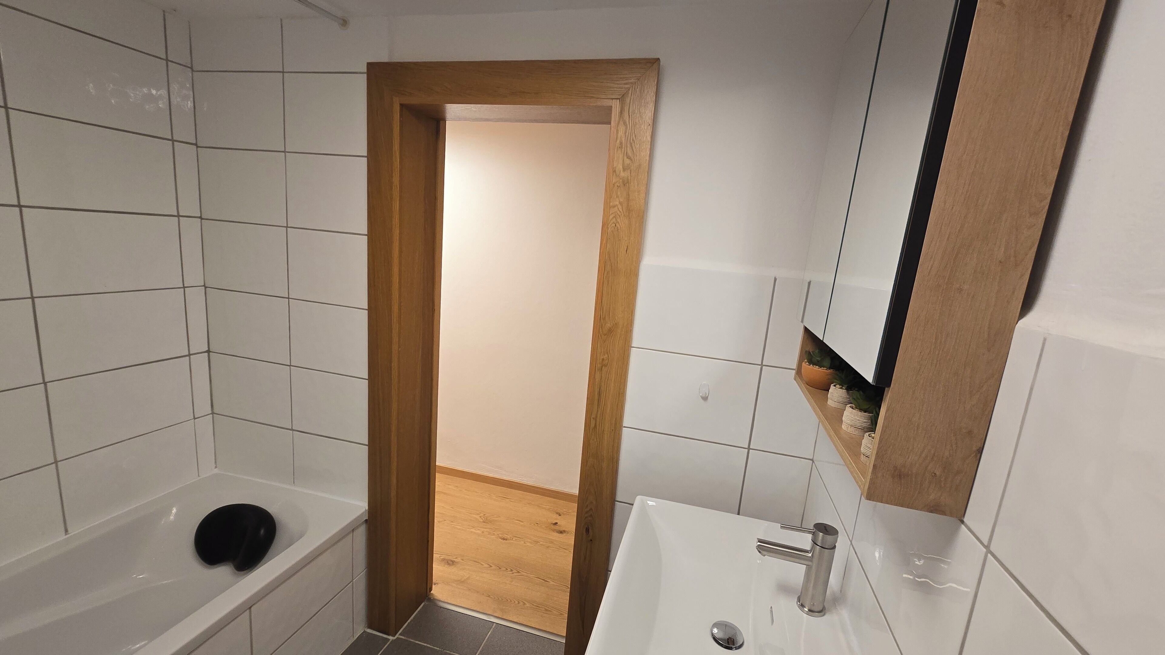 Ein Badezimmer mit Holztür, weißen Fliesen, einer Badewanne, einem Waschbecken mit Spiegel und einem Regal mit Pflanzen.