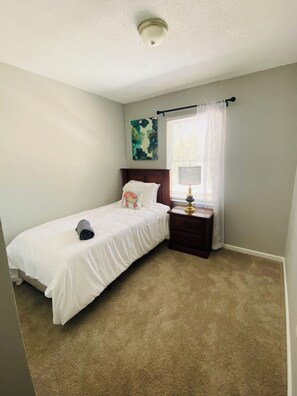 Une chambre à coucher avec un lit, une table de chevet, une lampe, une fenêtre avec des rideaux et un luminaire au plafond.