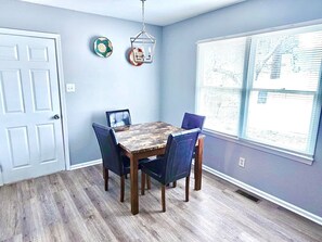 Un coin repas avec une table en bois et quatre chaises, une porte blanche et une fenêtre avec des stores.