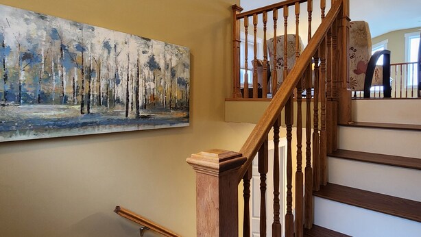 A wooden staircase with a railing, a painting of trees on the wall, and a patterned chair in the background. - Pilley's Perch - Entire home in Pilley's Island, Canada. Beautiful Ocean View (Pilley's Island)