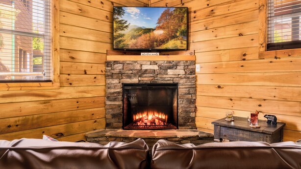 Interior acogedor de una cabaña con chimenea de piedra, asientos de cuero y un televisor montado en la pared.